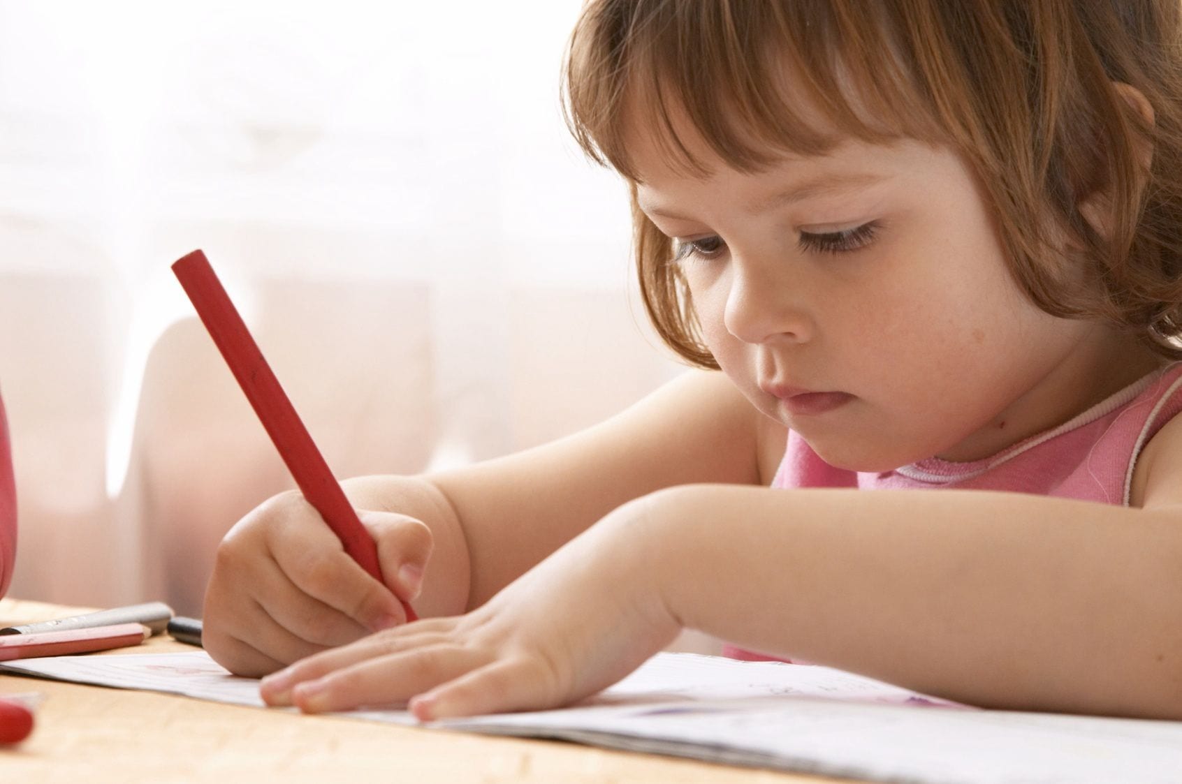 A little girl is writing with a red pencil.